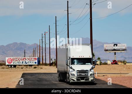 Il semi-camion bianco con rimorchio a cassoni guida lungo una strada desertica negli Stati Uniti occidentali, con vista frontale centrata con pali, cartelloni e montagne lontane. Foto Stock