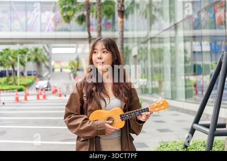 Giovane donna persona felice viaggio turista ama la musica delle vacanze, giocare ukulele all'aperto sulla vivace strada urbana della città, abbracciare lo stile di vita estivo con j Foto Stock