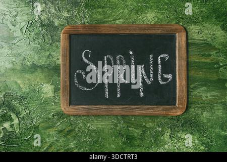 Le lettere a mano con iscrizione calligrafica si aprono su una lavagna nera vintage su sfondo verde. Piatto, spazio. Il concetto di primavera in arrivo. Foto Stock