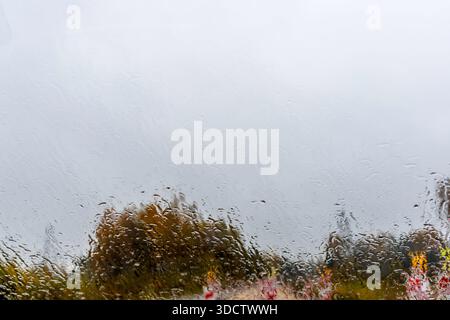 Guarda attraverso un parabrezza bagnato coperto da gocce di pioggia, mostrando un cantiere stradale sfocato con barriere rosse e bianche in una giornata cupa. Foto Stock