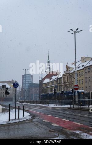 Le nevicate soffici coprono Wroclaw mentre un tram blu MPK passa davanti alle facciate, una guglia della chiesa e una torre di vetro distante. Asfalto bagnato e pista ciclabile rossa ma Foto Stock