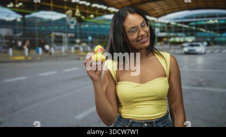 La donna tiene l'anatra di gomma vicino alla mano sollevata e alla spalla esposta sul marciapiede dell'aeroporto, sorridendo e inclinandosi leggermente in avanti; gioia giocosa. Foto Stock