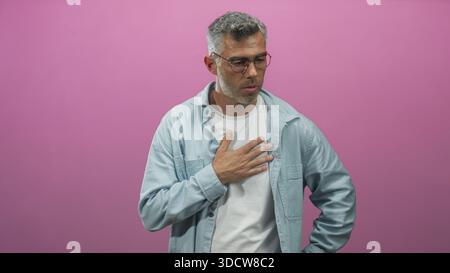 Uomo con la mano sul petto, gli occhi chiusi e la bocca leggermente aperta, che sospira in studio su uno sfondo rosa, indossa occhiali e una camicia azzurra; re Foto Stock