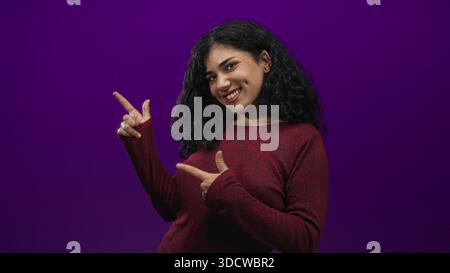 Giovane donna bruna sorridente e che punta entrambe le dita a lato in uno studio viola che indossa un maglione marrone; giocosa sicurezza. Foto Stock