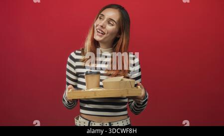 La donna tiene in mano un vassoio di legno con tazzina da asporto e scatole di cibo, sorridendo sullo sfondo di uno studio rosso; allegra ospitalità. Foto Stock