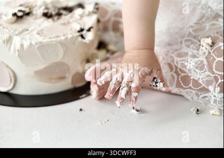 Un dolce primo piano di un bambino in un delicato abito in pizzo che sfonde gioiosamente una torta decorata con morbidi motivi floreali. Le consistenze cremose e la minuscola h Foto Stock