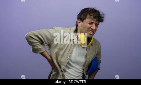 Uomo ispanico che indossa cuffie a cancellazione del rumore e tiene in mano un elmetto soffre con uno sfondo viola. Foto Stock