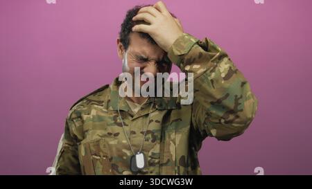 Il giovane soldato ispanico in uniforme mimetica tiene in mano le targhette per cani d'argento e preme mano sulla fronte in studio rosa; stress. Foto Stock