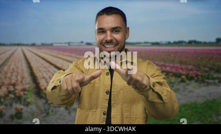 Uomo sorridente mentre punta entrambe le dita verso la fotocamera tra i tulipani in fiore in una foresta; giocosa sicurezza. Foto Stock