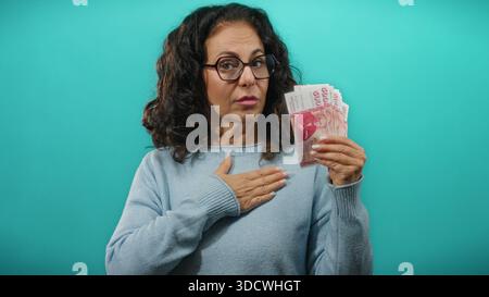 Donna di mezza età che tiene in mano banconote della corona islandese mentre alza la mano con un gesto di giuramento su uno sfondo blu. Foto Stock