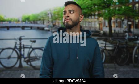 Uomo con le mani sulle guance per il mal di denti sulla strada del canale di amsterdam vicino alle biciclette e al ponte, con cappuccio, un aspetto doloroso; preoccupazione urbana. Foto Stock