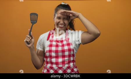 Donna che indossa grembiule a quadri rosso e bianco e maglietta bianca con spatola con occhi che ombreggiano le mani in studio arancione; divertiti con i momenti di cucina. Foto Stock