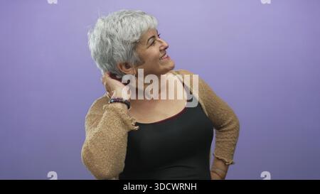 Donna senior taglia che tocca i capelli con la mano dietro il collo, sorride e guarda in alto, indossa braccialetto e cardigan in studio; gioiosa fiducia. Foto Stock
