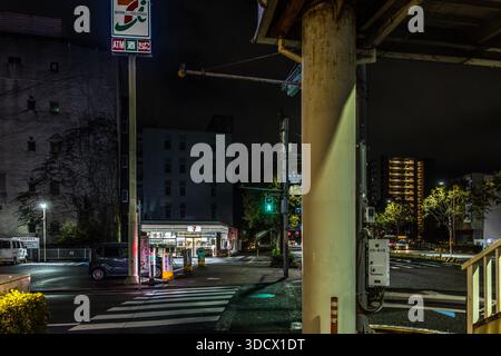 L'insegna illuminata di un negozio 7-Eleven illumina un incrocio notturno a Wakayama, Giappone, con un segnale pedonale verde. Un'unica auto è parcheggiata sul ciglio della strada davanti al negozio. Foto Stock