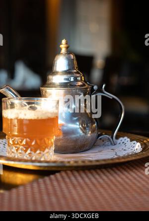 Tradizionale tè alla menta marocchino servito in una teiera decorativa in metallo e bicchiere su un vassoio Luce calda, atmosfera autentica e ospitalità culturale conc Foto Stock