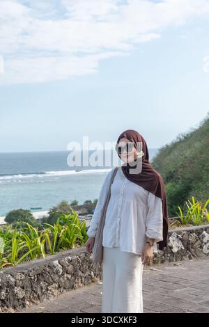 Donna musulmana che indossa un hijab in piedi vicino a una scogliera costiera con vista sull'oceano, che rappresenta lo stile di vita dei viaggi e la Moda Moda nella natura Foto Stock