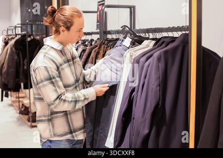 Giovane con acconciatura di panini che sceglie pantaloni blu in un negozio di abbigliamento mentre acquista nuovi capi di abbigliamento. Foto di alta qualità Foto Stock