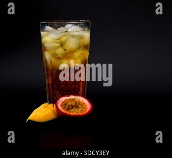 Bicchiere alto di succo di frutta con ghiaccio su sfondo nero, pezzi di frutto della passione maturo e mango nelle vicinanze. Primo piano. Foto Stock