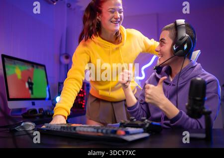 Due donne che indossano le cuffie che giocano insieme a un videogioco in una sala giochi colorata. Sono in possesso di controller di gioco, interagiscono con lo schermo e si divertono in una sessione multigiocatore entusiasmante || Model rilasciato Foto Stock