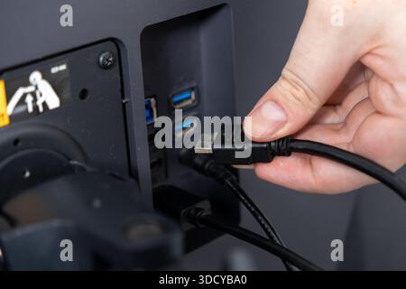Primo piano di una mano umana inserendo con attenzione un cavo HDMI nel retro di un monitor, evidenziando la configurazione dei dispositivi elettronici e la perfetta integrazione digitale Foto Stock