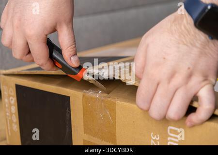 Primo piano di mani umane aprendo con attenzione una confezione di cartone marrone con nastro e un coltello, che rappresenta la consegna del prodotto e la nuova merce Foto Stock