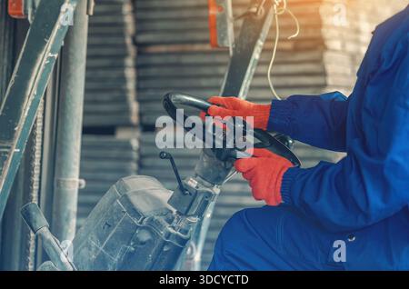 Donna in abbigliamento da lavoro blu che guida carrelli elevatori all'interno di un magazzino industriale. Lavoratore professionista che opera macchinari pesanti in ambito logistico o produttivo Foto Stock
