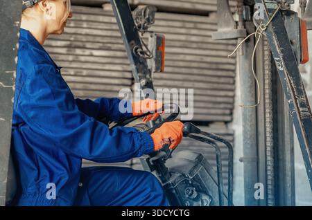 Donna in abbigliamento da lavoro blu che guida carrelli elevatori all'interno di un magazzino industriale. Lavoratore professionista che opera macchinari pesanti in ambito logistico o produttivo Foto Stock