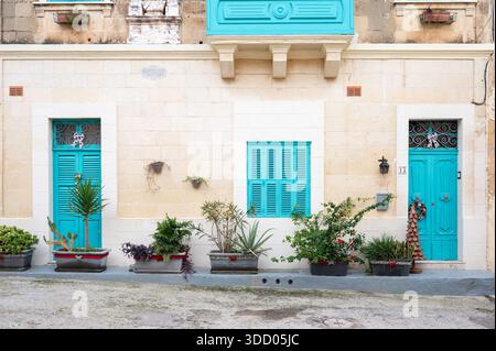 Stradine strette fiancheggiate da case semplici caratterizzate da porte e finestre in legno colorato adagiate in pietra calcarea ocra, che riflettono il fascino modesto di una tradizione Foto Stock