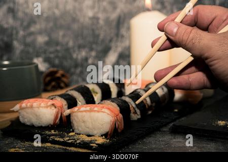 Utilizza a mano bacchette di legno per prendere il sushi da una tavola di ardesia con nigiri e panini, concetto di assaggiare la cucina tradizionale giapponese a casa. Foto Stock