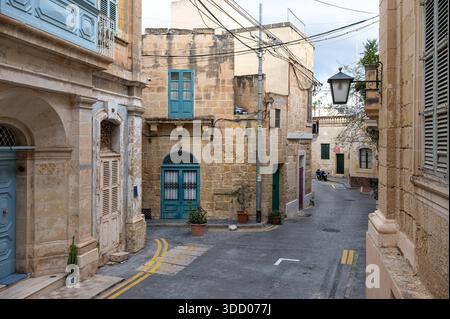 Stradine strette fiancheggiate da case semplici caratterizzate da porte e finestre in legno colorato adagiate in pietra calcarea ocra, che riflettono il fascino modesto di una tradizionale città maltese di Iz-Zeytun, Malta, 10 dicembre 2025. Foto Stock