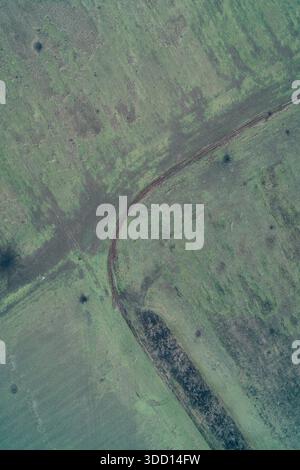 Vista dall'alto verticale della strada sterrata che si snoda attraverso il campo erboso. Ripresa con droni ad alta risoluzione di percorsi rurali, tracce di pneumatici sul terreno e sfondo con texture verde crudo con spazio di copia. Foto Stock
