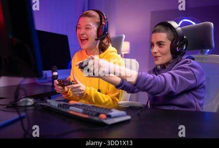 Due donne che indossano le cuffie che giocano insieme a un videogioco in una sala giochi colorata. Sono in possesso di controller di gioco, interagiscono con lo schermo e si divertono in una sessione multigiocatore entusiasmante || Model rilasciato Foto Stock