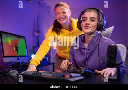 Due donne che indossano le cuffie che giocano insieme a un videogioco in una sala giochi colorata. Sono in possesso di controller di gioco, interagiscono con lo schermo e si divertono in una sessione multigiocatore entusiasmante || Model rilasciato Foto Stock