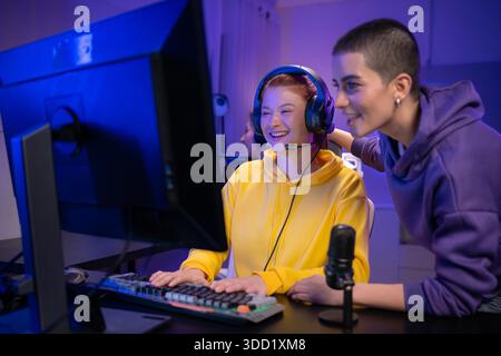 Due donne che indossano le cuffie che giocano insieme a un videogioco in una sala giochi colorata. Sono in possesso di controller di gioco, interagiscono con lo schermo e si divertono in una sessione multigiocatore entusiasmante || Model rilasciato Foto Stock