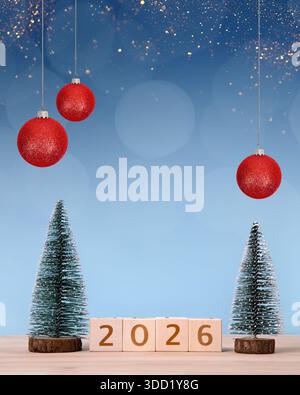 Capodanno 2026 e cartolina di Natale. Cubetti di legno con numeri 2026, palle rosse e alberi di Natale verdi su un tavolo di legno e sfondo blu. Foto Stock