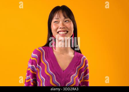 Bellissimo ritratto di donna cinese su sfondo colorato. Giovane donna asiatica che posa in studio, indossa un maglione viola vibrante e un fulmine rosa Foto Stock