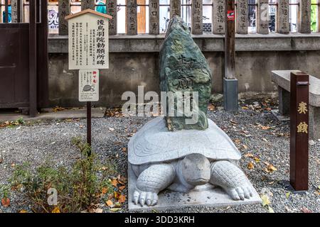 Una tartaruga di pietra porta la pietra commemorativa „Chokuganjo“ al santuario Mitsu Hachiman (Mitsu-gu) a Osaka, in Giappone. Riporta che il santuario funse da luogo di preghiera imperiale per l’imperatore abdicato Go-Toba nel periodo Kamakura. Il cartello accanto invita i visitatori ad accarezzare la tartaruga e a esprimere un desiderio. La tradizione fa risalire il santuario al 749, quando un santuario portatile di Usa approdò qui. I bombardamenti su Osaka distrussero tutti gli edifici nel 1945; furono ricostruiti nel 1960. Foto Stock