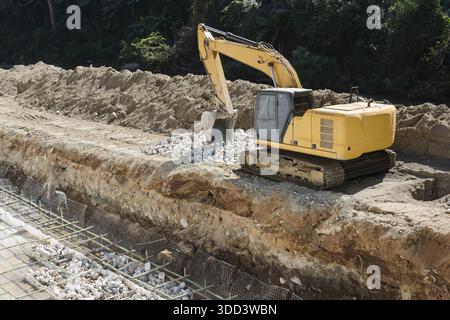 Macchina da escavatore pesante di grandi dimensioni gialla che sposta terra e terreno di scavo in cantieri industriali affollati vicino a strutture in cemento e foreste durante il periodo di Foto Stock