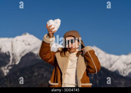Una donna con il cappotto marrone e gli occhiali da sole tiene una palla di neve a forma di cuore sullo sfondo di montagne innevate sotto un cielo azzurro. Foto Stock
