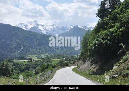 Una strada tortuosa serpeggia attraverso lussureggianti colline verdi sotto un cielo blu luminoso con nuvole sparse. Le montagne innevate si innalzano maestosamente in lontananza Foto Stock