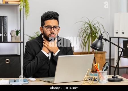 Uomo d'affari indiano libero professionista che si sente male malato malato affetto da mal di gola ferito deglutizione dolore tocca il collo lavorare a casa ufficio sul posto di lavoro. Maschio indù in tuta formale ha un raffreddore, influenza, virus Foto Stock