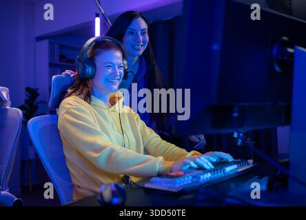 Due donne che indossano le cuffie che giocano insieme a un videogioco in una sala giochi colorata. Sono in possesso di controller di gioco, interagiscono con lo schermo e si divertono in una sessione multigiocatore entusiasmante || Model rilasciato Foto Stock