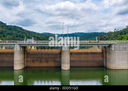 Una diga di cemento si estende su acque verdi calme, adagiate su uno sfondo di fitta foresta tropicale e colline ondulate sotto un cielo parzialmente nuvoloso. L'immagine evidenzia forme ingegneristiche, superfici riflettenti e il contrasto tra infrastruttura e paesaggio naturale. Foto Stock