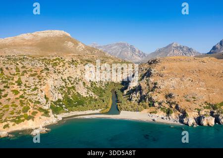 Immagine aerea di uno stretto fiume che si snoda attraverso un'aspra valle di montagna e una spiaggia di sabbia vuota a Preveli, Creta. Il paesaggio presenta aridi pendii rocciosi, vegetazione sparsa e un mare blu profondo sotto un cielo senza nuvole. Foto Stock
