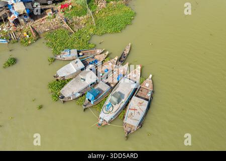 Vista aerea di diverse barche in legno con ponti coperti ancorati lungo la riva verde del fiume a Chau Doc, Vietnam. L'immagine evidenzia i vasi galleggianti, le piante acquatiche e la superficie dell'acqua strutturata del delta del Mekong. Foto Stock