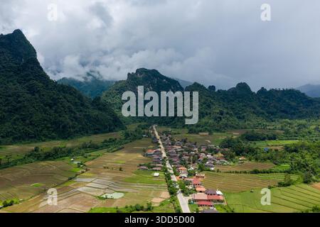 Vista aerea di un villaggio rurale annidato tra ripide montagne verdi e campi di riso patchwork nel Laos settentrionale. Le spettacolari nuvole si stendono in basso sulle colline carsiche, con un'unica strada che conduce attraverso la vibrante terra agricola e la fitta foresta. Foto Stock