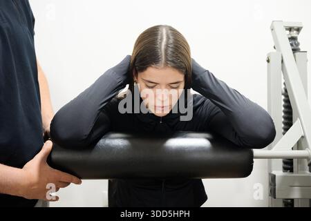 Personal trainer che assiste una giovane donna nell'esercizio di allungamento assistito della schiena sulla macchina per il fitness per la forza e la riabilitazione Foto Stock