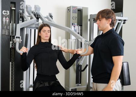 Una giovane donna che lavora sui muscoli toracici con una macchina da palestra, ricevendo la guida di un personal trainer maschile Foto Stock