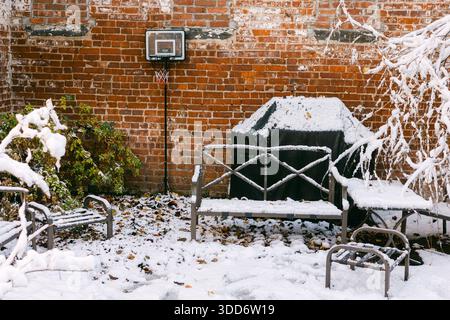 Coperte di neve un cortile con sedie, un grill e un canestro da basket contro una parete di mattoni in inverno. Foto Stock