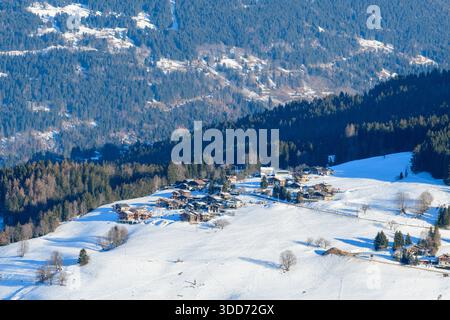 Un gruppo di chalet alpini tradizionali si trova su una collina illuminata dal sole e coperta di neve vicino a Megeve, delimitata da fitte foreste sempreverdi e da ondulate montagne. Foto Stock